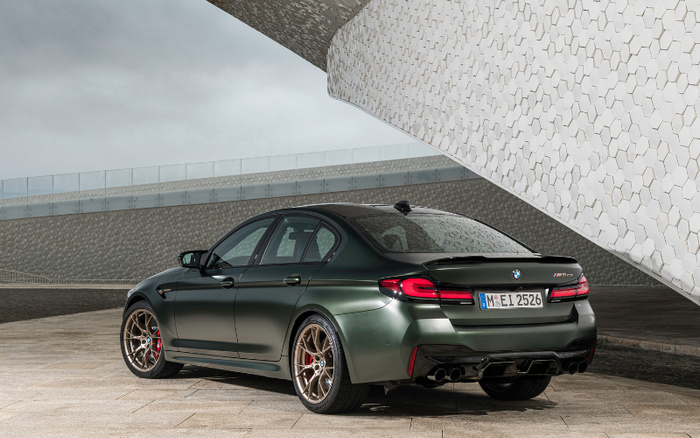 Rear and side view of a dark green BMW M5 CS parked on a paved area under a modern architectural structure with hexagonal patterns. The car features red brake calipers, gold alloy wheels, and a visible licence plate reading 'M EI 2526'. The setting is outdoors with cloudy skies, showcasing a sleek, high-performance vehicle suitable for those interested in used cars and new cars.