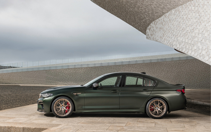 Side view of a dark green luxury sedan parked on a stone pavement beside a modern architectural building with curved, textured walls under a cloudy sky. The car is a new model with sporty alloy wheels and red brake calipers, showcasing sleek design and premium features suitable for used cars and new cars market.