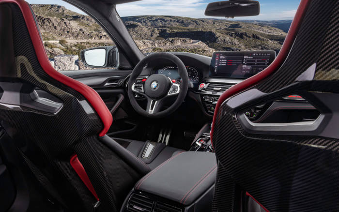 Interior view of a BMW car featuring sporty black carbon fibre bucket seats with red accents, a multifunctional steering wheel with the BMW logo, and a digital dashboard displaying various vehicle settings. The centre console includes a large touchscreen showing media options and car data. The background reveals a scenic rocky mountain landscape visible through the windscreen and side window. This image highlights the luxurious and modern design typical of new cars, relevant for used cars and new cars markets.