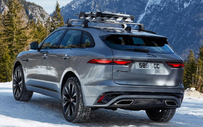 Silver Jaguar F-PACE SUV with roof rack parked on snow-covered ground in a mountainous area with coniferous trees and steep rocky peaks in the background, showing rear and side view with license plate number 589 SCC, suitable for advertisements of used cars and new cars.