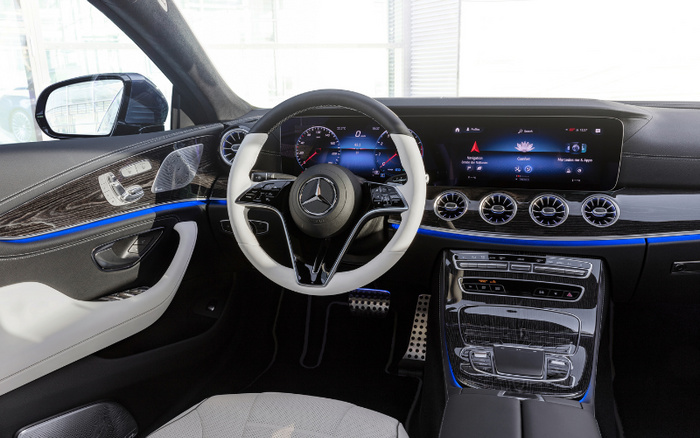 Interior view of a luxury car showcasing the steering wheel with the Mercedes-Benz logo, a digital dashboard displaying speedometer and various car information, and an infotainment screen with features like navigation, media, and Mercedes me apps. The car interior features black and cream leather seats, blue ambient lighting, modern air vents, and a wooden trim finish. Perfect example of new cars and used cars offering advanced technology and stylish design.