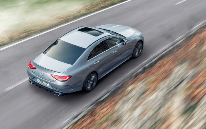 Silver-grey Mercedes-Benz sedan with tinted windows and sunroof driving on a smooth paved road surrounded by blurred greenery and autumn leaves, representing luxury used cars and new cars.