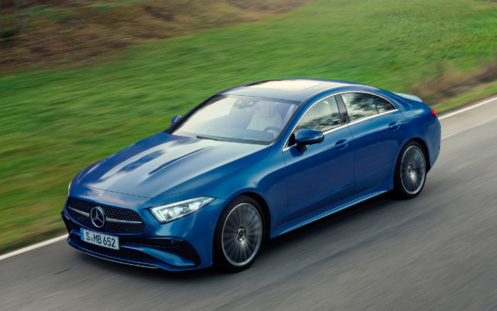Blue Mercedes-Benz CLS 2024 model driving on a road next to green grass, showcasing sleek design and modern headlights. Licence plate reads S MB 652. Suitable for used cars and new cars buyers interested in luxury vehicles.