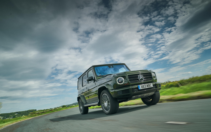 A dark green Mercedes-Benz G-Class SUV driving on a rural road under a partly cloudy sky during daytime, with the license plate reading '463 MBC'. The image showcases a luxury new car, suitable for promoting both used cars and new cars in automotive sales.