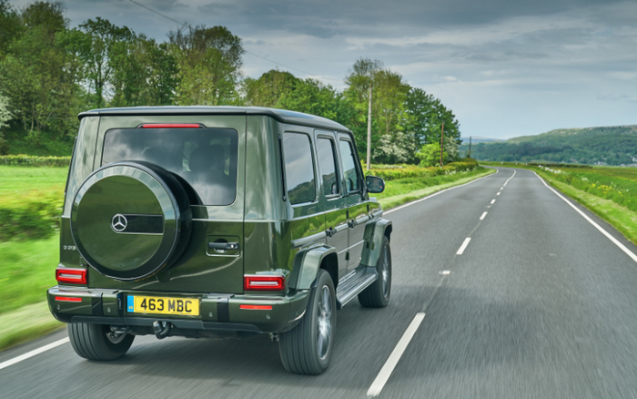 Green Mercedes-Benz G-Class SUV with licence plate 463 MBC driving on a two-lane road through a rural landscape with green fields and trees under a cloudy sky, representing used cars and new cars on sale.