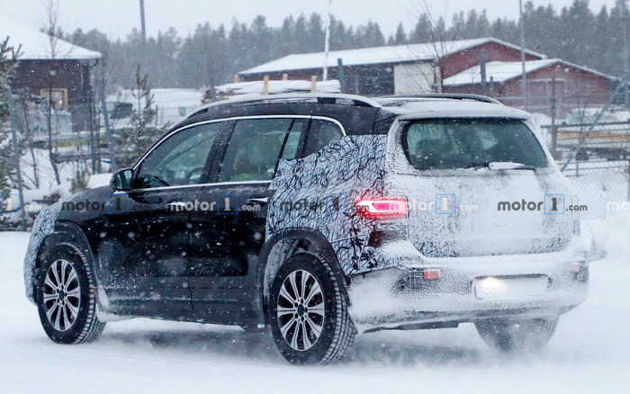 Black and white camouflaged prototype SUV driving on a snowy road with snow-covered ground and buildings in the background. The rear and part of the side of the car are covered in testing camouflage wrap, with rear lights illuminated. The image has watermarked text 'motor1.com' repeatedly over the scene, indicating it is likely a spy shot of a new car model in winter testing conditions. Keywords: used cars, new cars.