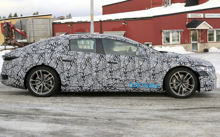 Side view of a new car prototype covered in black and white camouflage wrap with intricate patterns, parked on a snowy road in front of red industrial buildings. The car has sporty alloy wheels and the word 'carnow' written in blue on the lower side panel. The background shows snow-covered roofs and a sign reading 'SWEROCK TRANSPORT & MAS...'. This image relates to new cars and used cars in the context of vehicle testing and development.