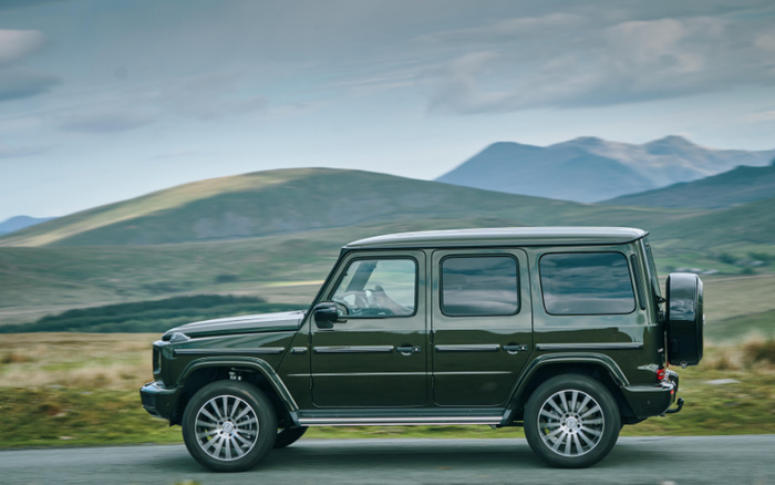 Dark green luxury SUV driving on a road with green hills and mountains in the background, representing a used car or new car for sale in a scenic countryside setting.