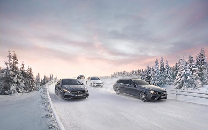 Four Mercedes-Benz cars driving on a snow-covered road surrounded by snow-covered pine trees under a pastel pink and blue sky at dusk. The cars include black and white models, showcasing sleek designs and winter performance. This image highlights both used cars and new cars suitable for winter driving conditions.