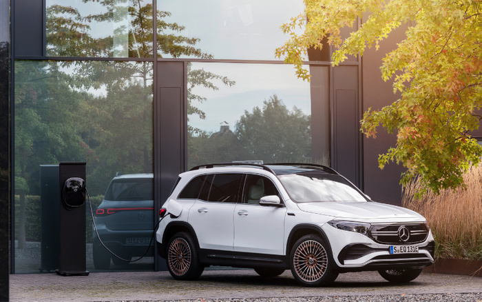 White Mercedes-Benz electric SUV charging at a modern home charging station with large glass windows reflecting the vehicle. The license plate reads 'S EQ 131E'. Surrounding autumn foliage and a paved driveway visible, showcasing a new car model. Keywords: used cars, new cars.