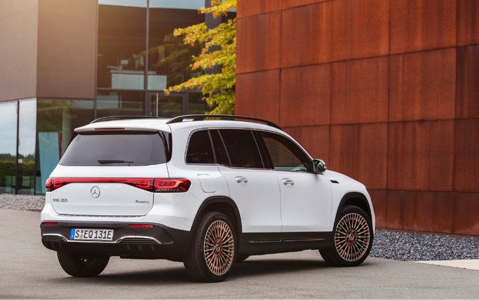 Rear and side view of a white Mercedes-Benz EQB 350 4MATIC electric SUV parked outdoors next to a building with rusty brown panel walls and glass windows. The car features sleek taillights, a roof rack, and stylish multi-spoke alloy wheels. The licence plate reads S EQ 131E. The scene suggests a modern, urban environment suitable for showcasing used cars and new cars.