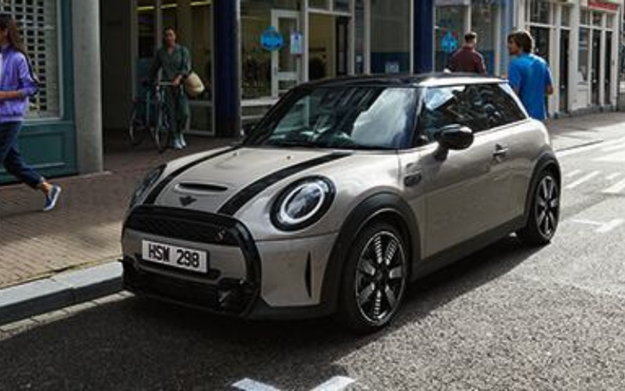 Silver Mini Cooper with black racing stripes parked on a city street with licence plate HSM 298. Pedestrians walk nearby past a shop with glass windows and a cyclist is in the background. Keywords used cars and new cars.