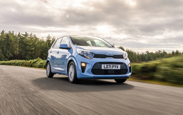 Light blue Kia hatchback car with registration plate LD71 PYV driving on a road with greenery and trees on the side under a cloudy sky, representing used cars and new cars.
