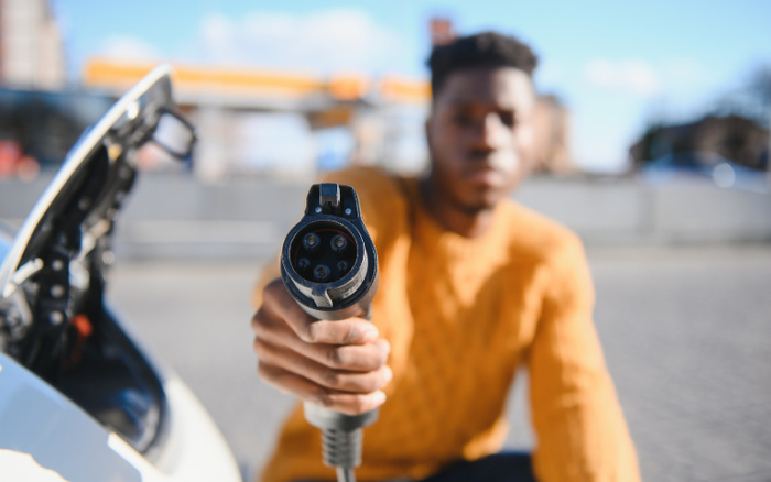 Close-up of a person in a mustard yellow sweater holding an electric car charging plug towards the camera next to a white electric vehicle, representing modern technology for both used cars and new cars.