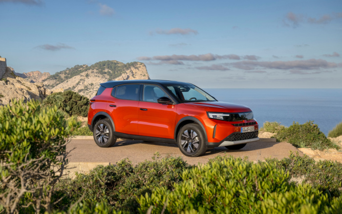 Bright orange new car parked on a scenic coastal road with cliffs and the sea in the background, surrounded by green bushes and clear blue sky, ideal for showcasing new cars or used cars.
