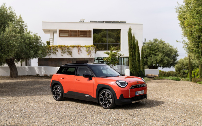 Bright orange compact crossover car parked on gravel driveway in front of a modern white house with large windows and surrounded by trees. The car has a black roof and detailing, stylish alloy wheels, and a European license plate. The image relates to used cars and new cars available for purchase.