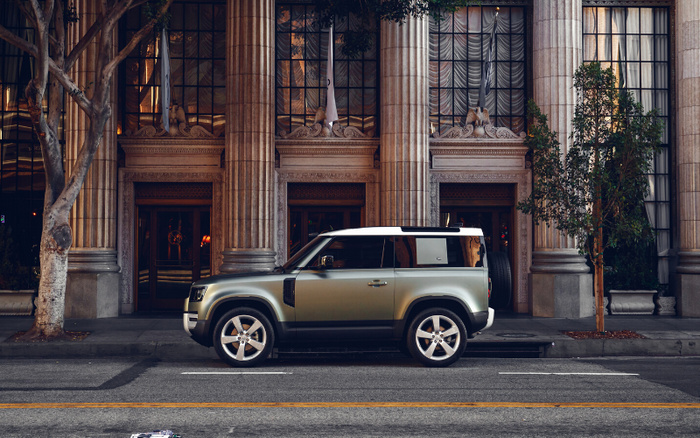 Side view of a metallic green and white Land Rover Defender parked in front of a grand classical building with tall columns and large windows, illustrating a new car model in an urban setting with trees and reflections in the windows, suitable for advertising used cars and new cars.