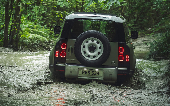 Rear view of a dark green Land Rover Defender 4x4 with license plate PBS 534 driving through a shallow, muddy water stream in a dense forest, highlighting its off-road capabilities, suitable for buyers interested in robust used cars and new cars.