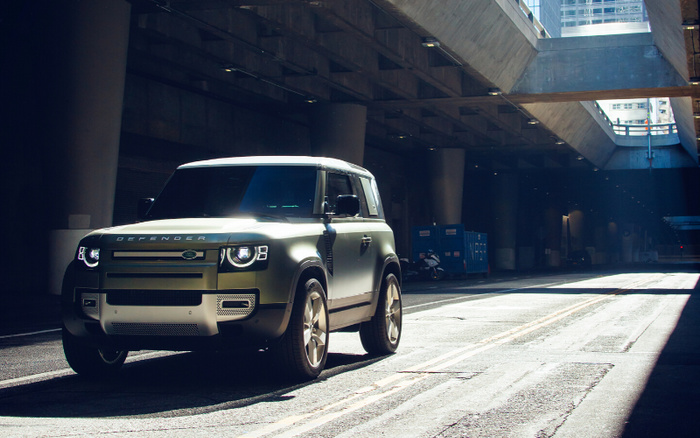 A matte green Land Rover Defender parked under a city bridge or overpass, with sunlight casting shadows on the road. The vehicle is positioned on the left side of the image, highlighting the front and side profile including the distinctive 'DEFENDER' lettering on the bonnet. The urban setting features concrete pillars and structural beams above, with light filtering through gaps. This image is suitable for showcasing used cars or new cars in an urban environment.