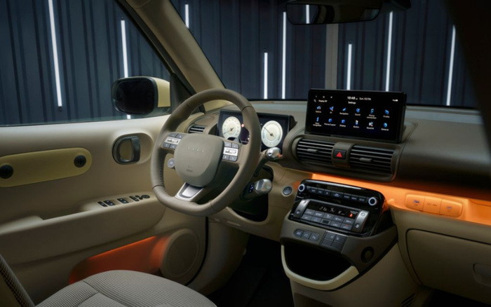 Interior view of a modern car displaying the dashboard and steering wheel, with digital instrument cluster showing speedometer and other gauges, and a touchscreen infotainment system displaying options such as phone, navigation, radio, media, and settings. The car features beige and orange trim with various control buttons on the steering wheel and centre console, illustrating the cabin design of new cars at a dealership that offers used cars and new cars.