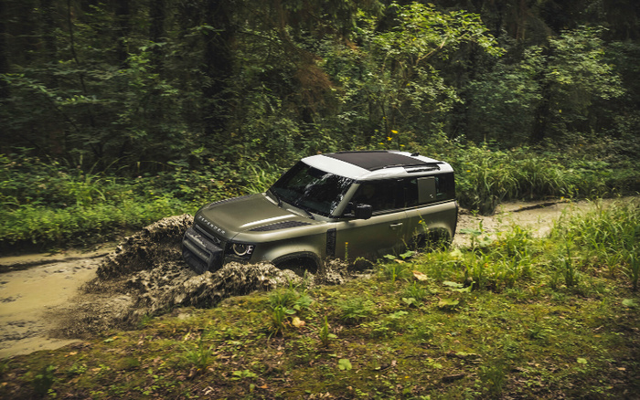 A green Land Rover Defender driving through a muddy off-road trail surrounded by dense green forest, showcasing its off-road capabilities. The vehicle is splashing mud as it navigates the terrain, highlighting durability and ruggedness, relevant for both used cars and new cars enthusiasts seeking adventure-ready vehicles.