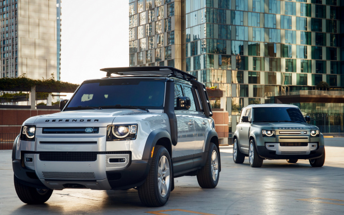 Two silver Land Rover Defender SUVs parked in an urban setting with tall glass buildings in the background, showcasing modern design and rugged features, suitable for marketing used cars and new cars.