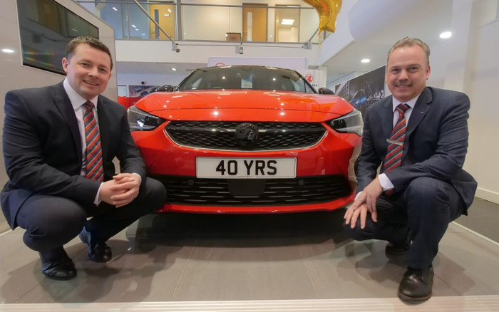 Two men in business suits and striped ties crouching beside a red Vauxhall car with the personalised number plate '40 YRS' inside a showroom, representing a celebration of 40 years, relevant to used cars and new cars sales and dealership services.