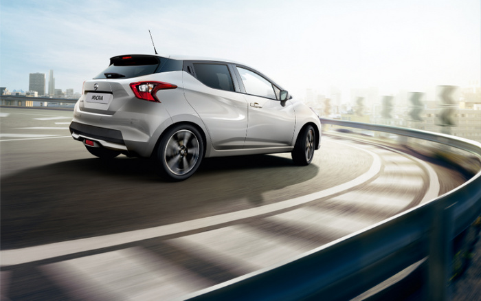 Silver Nissan Micra compact car cornering on an elevated urban road with city skyline in the background. Used cars and new cars concept featuring a modern hatchback with sleek design and alloy wheels.