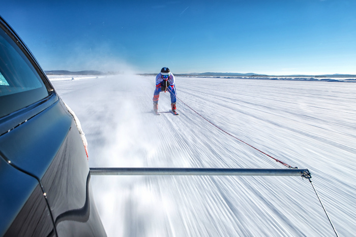 A person wearing winter sports gear and helmet is ski racing, being towed behind a black car on a snowy, icy landscape under a clear blue sky, illustrating the use of used cars or new cars in extreme winter conditions.
