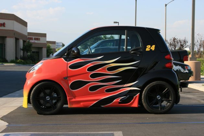 Side view of a compact two-door Smart car with a customised flame paint design in red, black, yellow and white. The car is parked in a parking lot with the number 24 in yellow on the upper rear side panel. Background includes a commercial building and clear sky. Ideal image for showcasing used cars or new cars with unique customisation.