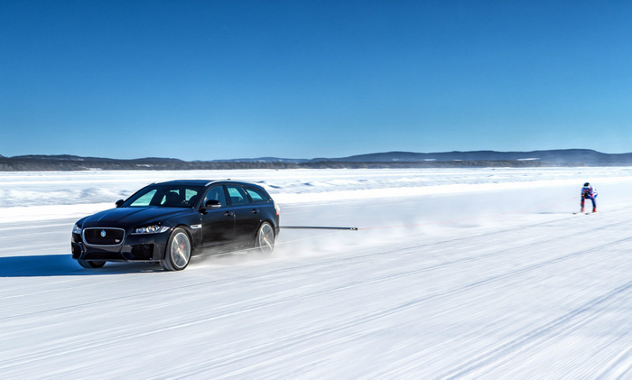 Black Jaguar XF Sportbrake towing a skier on a snowy frozen lake under clear blue sky with distant hills, showcasing a new car in an outdoor winter testing or demonstration environment, relevant for used cars and new cars.