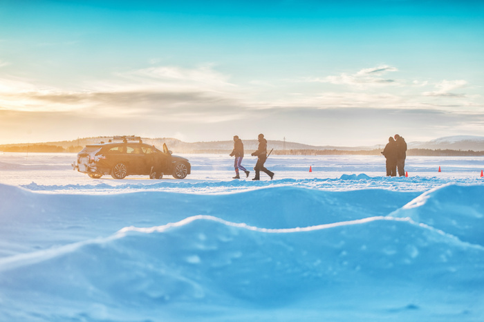 A black car covered in snow parked on a snowy surface at sunset with people walking nearby and orange cones marking the area, suggesting winter testing or driving conditions involving used cars and new cars.