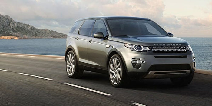 Silver Land Rover Discovery driving on a coastal highway with the sea and rocky cliffs in the background, showcasing a sleek design of a used cars or new cars SUV.