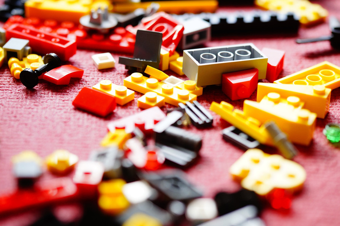 Close-up image of scattered LEGO bricks in red, yellow, black, grey, and white colours on a textured red surface, showing various shapes and sizes of the bricks.