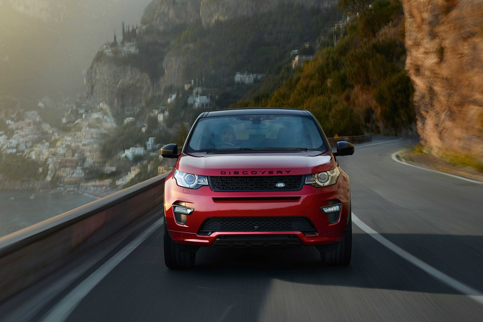Front view of a red Land Rover Discovery driving on a winding coastal road with mountainous terrain and buildings in the background, showcasing a new cars model ideal for used cars buyers seeking adventure and style.