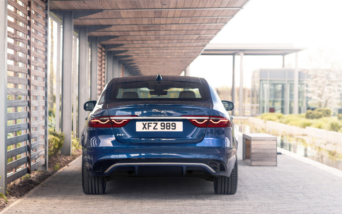 Rear view of a blue Jaguar XE car with vehicle registration plate XFZ 989 parked under a covered outdoor area with wooden slats and concrete pavement, highlighting details suitable for used cars and new cars listings.