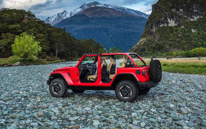 Red Jeep Rubicon with no doors on rocky riverbed surrounded by green trees and tall mountains with snow-capped peaks under a cloudy sky, showcasing an adventurous off-road vehicle ideal for used cars and new cars buyers seeking rugged performance.