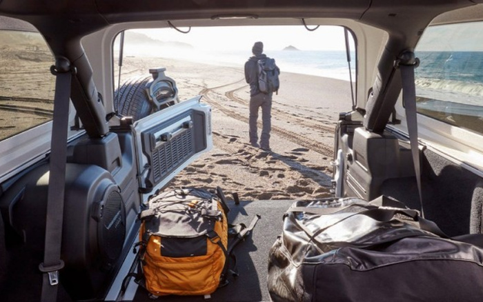 View from the rear interior of an off-road vehicle parked on a sandy beach. The back door is open, revealing two backpacks inside—one orange and gray, the other black. Outside, a person wearing a backpack stands near the shoreline looking at the sea. The scene evokes adventure and travel, highlighting the spacious cargo area suitable for luggage in a new or used car designed for outdoor use.
