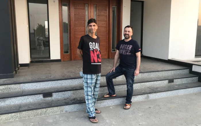 Two men standing outside a modern building with large wooden doors. One man is wearing a black t-shirt with the text 'GOAL N3' in red and white and blue checkered pants, while the other man is wearing a dark t-shirt with the text 'HIGH POWER' and dark jeans. Both are wearing sandals. The image does not depict used cars or new cars, but features casual attire and a contemporary entrance.