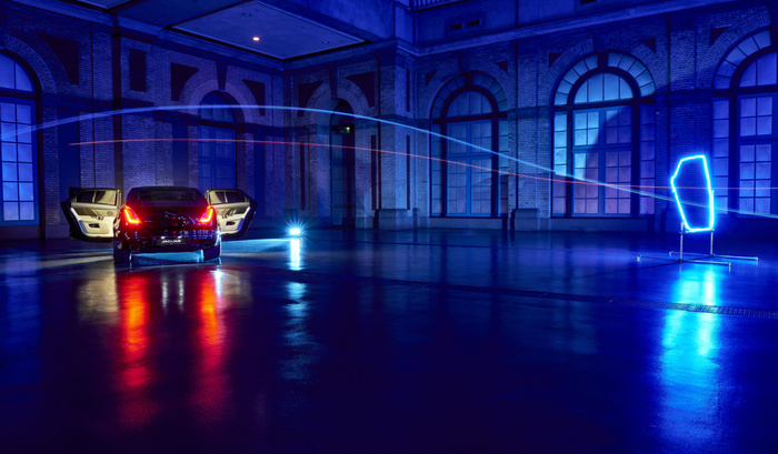 Rear view of a black Jaguar car with both rear doors open inside a large industrial building with tall arched windows and brick walls at night, illuminated by blue and red lights reflecting on the shiny floor, with a neon light installation on the right side and light trails crossing above the car, suitable for showcasing new cars and used cars in a stylish, modern setting.