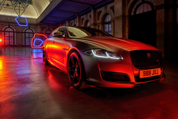 Sleek silver Jaguar luxury sedan illuminated by red and blue neon lights inside a large industrial-style building with arched windows. The car features distinctive LED headlights and black alloy wheels, with a visible license plate reading '888 JXJ'. The image showcases a stylish used car or new car in a dramatic, modern setting.