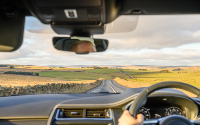 View from inside a car looking out over a long rural road with green fields and stone walls on either side. The image shows the dashboard, steering wheel, and rear-view mirror reflecting a partially visible driver. The scene captures the experience of driving, suitable for themes related to used cars and new cars.