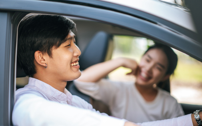 Two young people smiling and enjoying a drive in a car, one person in the driver's seat wearing a white shirt and an earring, the other person in the passenger seat blurred in the background; image suitable for topics on used cars and new cars.