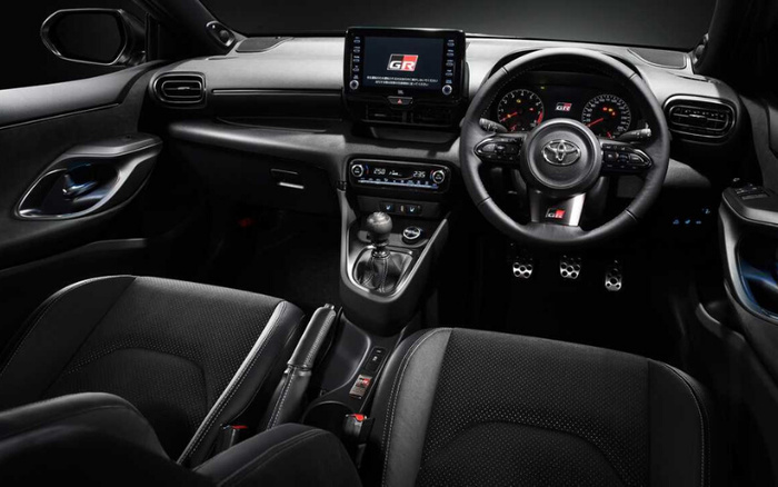 Interior view of a modern Toyota car showcasing the dashboard, steering wheel with GR logo, digital display screen showing 'Gazoo Racing GR', and manual gear shift. The image highlights features such as air vents, climate control panel with temperature settings, black fabric seats, and sporty metal pedals, representing both used cars and new cars options.