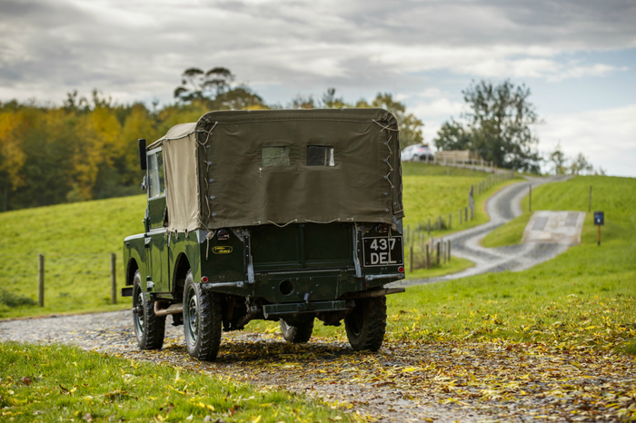 Rear view of a classic green Land Rover Series vehicle with canvas cover driving on a gravel country road, registration plate 437 DEL, surrounded by green fields and autumn trees under a cloudy sky, suitable for showcasing used cars and new cars in rural or off-road settings.