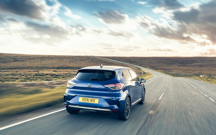 Rear view of a blue Renault Clio hatchback driving on a winding countryside road under a cloudy sky, showcasing a modern compact car suitable for both used cars and new cars buyers, with a visible yellow UK license plate reading REN 145.