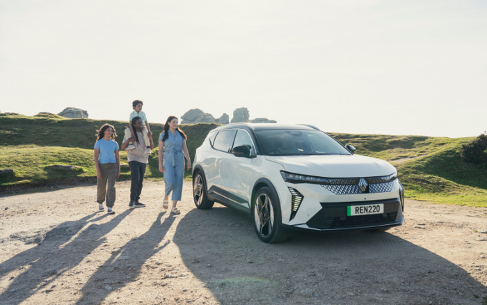 A white Renault SUV with license plate REN220 parked on a sunny gravel area with grassy hills in the background. A family of four walks beside the car, including a man carrying a child on his shoulders, a woman in a blue dress, and a young girl in casual clothes. The scene represents new cars suitable for family use, highlighting both used cars and new cars in a natural outdoor setting.