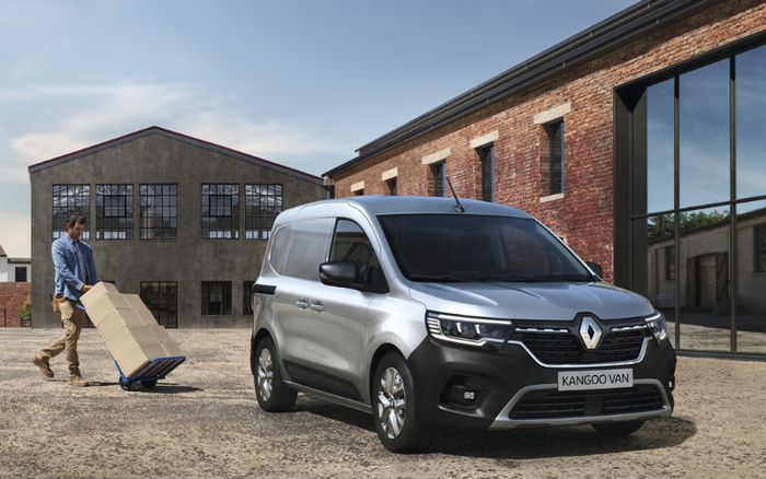 Silver Renault Kangoo Van parked in a courtyard with industrial-style brick and concrete buildings in the background. A man is pushing a hand truck with boxes behind the van. The van's front license plate reads 'KANGOO VAN'. This image highlights used cars and new cars suitable for commercial and delivery purposes.