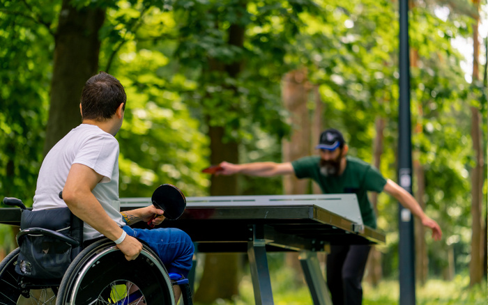 Two men playing table tennis outdoors in a park, one man in a wheelchair holding a paddle wearing a white t-shirt and blue pants, the other man in a green t-shirt and cap actively reaching to hit the ball, surrounded by green trees and natural light, no visible text related to used cars or new cars.