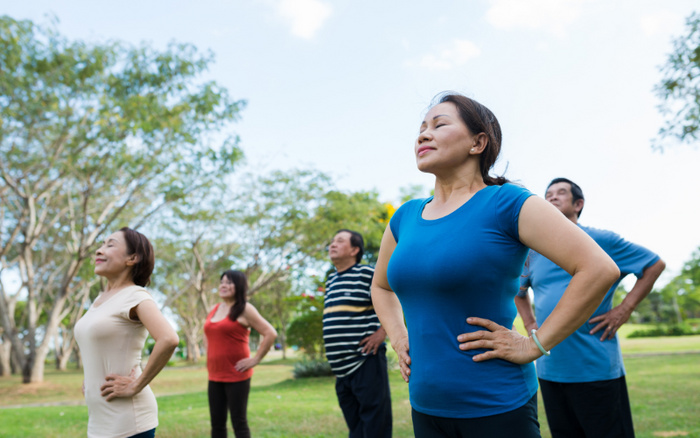 Group of middle-aged people standing outdoors in a park with hands on hips, eyes closed, and faces relaxed as they practise breathing or stretching exercises. The scene is bright with green trees and grass under a clear blue sky. This image does not feature used cars or new cars.